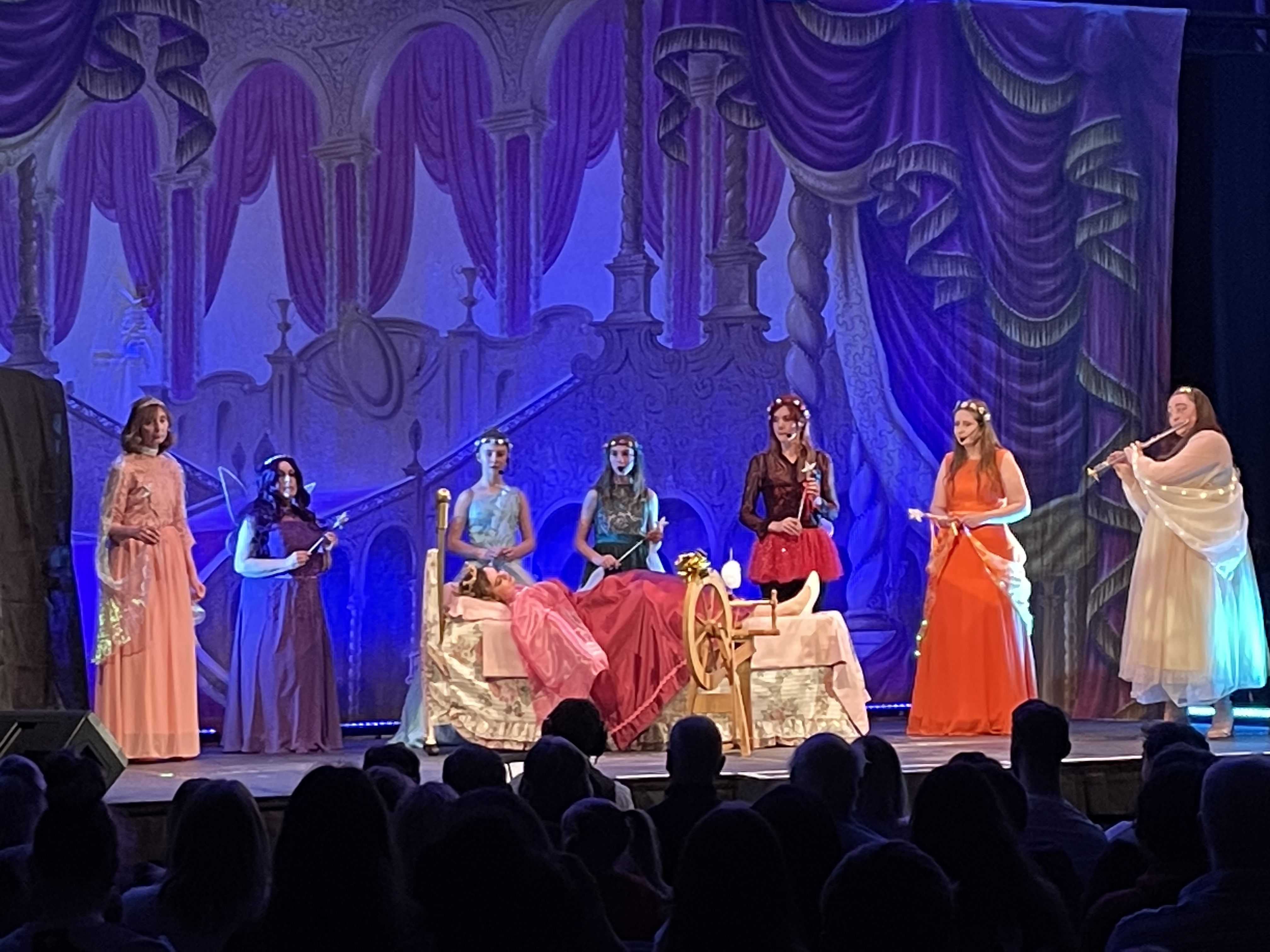 Seven colours, all the colours of the rainbow, surround The Sleeping Beauty, who rests peacefully on her bed. The spinning wheel that ahs caused her sleep is in the foreground. One fairy is playing a lullaby on her flute.